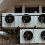 Couple in fashionable attire shares a kiss near industrial air conditioners, blending romance with urban grit.