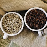 Overhead view of raw and roasted coffee beans in cups on burlap background.
